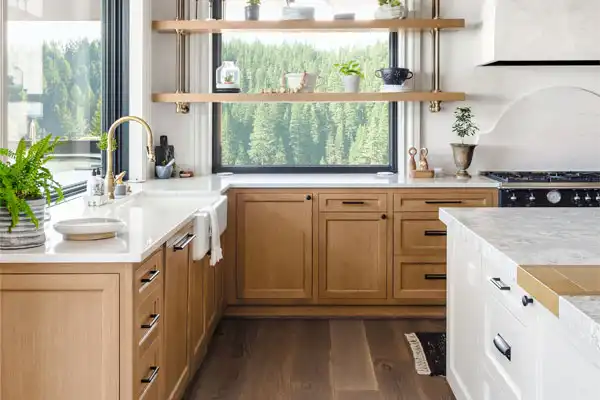 Modern kitchen with light wood cabinets, white countertops, a farmhouse sink, open shelves with plants, and a large window overlooking a forested landscape—perfect after a recent plumbing repair by a trusted plumber near me.