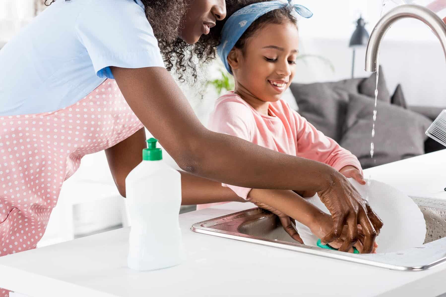 An adult and a child wash dishes together at a kitchen sink in their home, with water running from the tap—thanks to the best water heater—and a dish soap bottle on the counter.