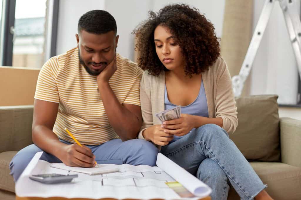 A man writes on paper beside a blueprint for a DIY HVAC project, while a woman next to him holds cash. Both are seated on a couch and appear focused on the task.