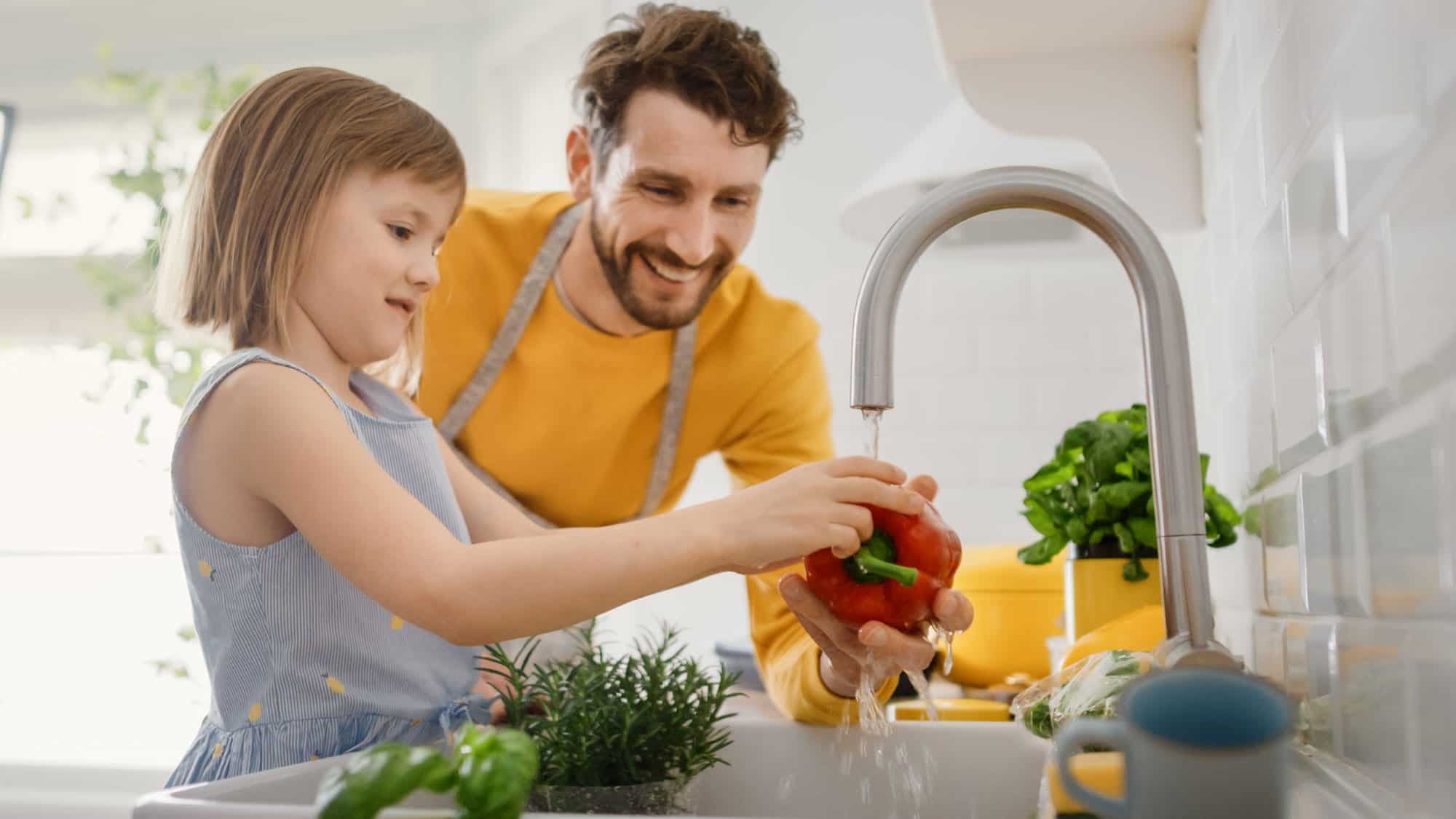 A man and a young girl wash a red bell pepper under a kitchen faucet, with fresh herbs and vegetables nearby on the counter, while sharing tips on proper garbage disposal maintenance.