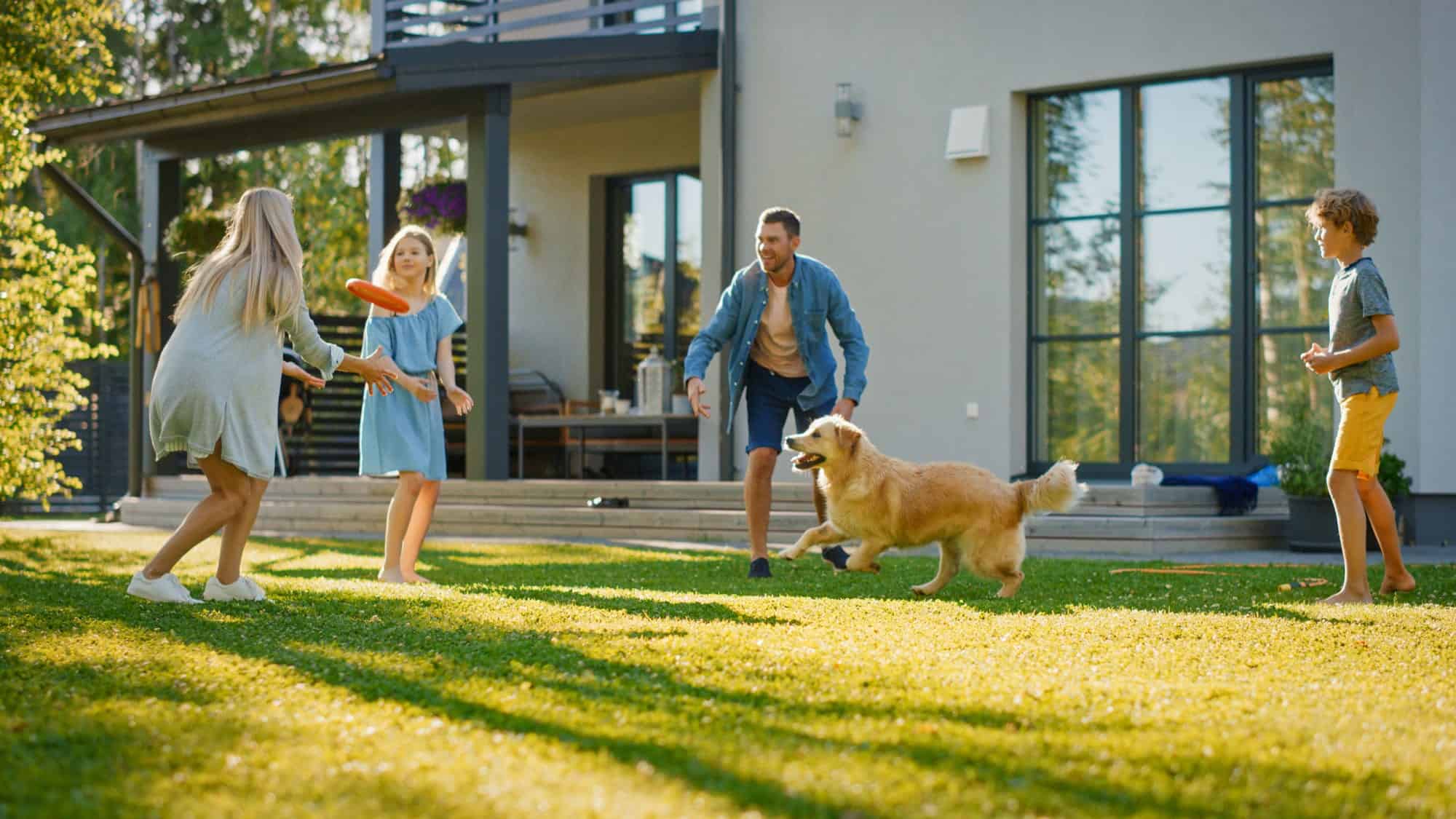 A family of four and a dog play with a frisbee on the lawn in front of a modern house with an advanced HVAC system that helps alleviate seasonal allergies on a sunny day.