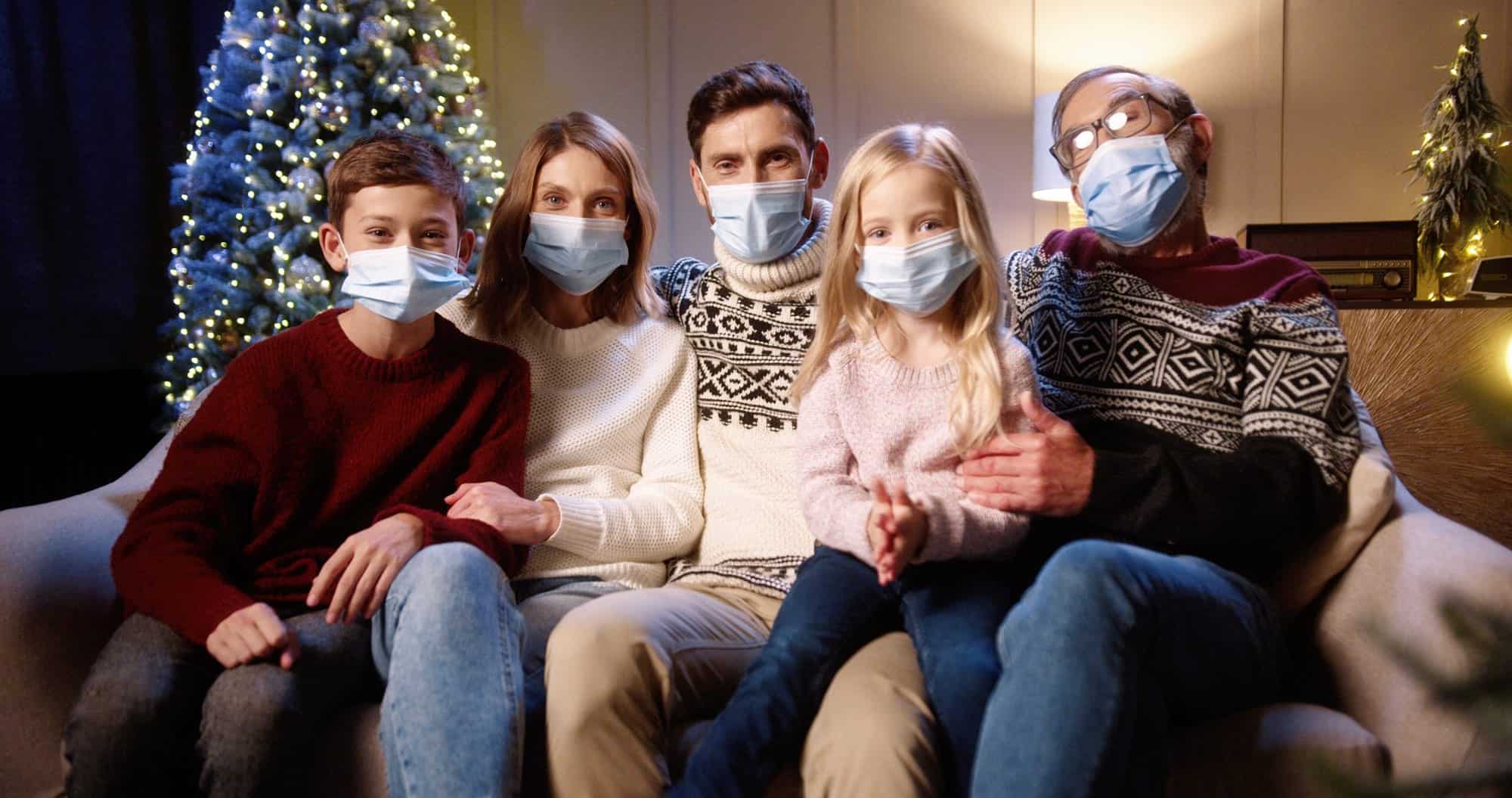 Five people wearing masks sit on a couch in front of a decorated Christmas tree, posing for a photo and practicing holiday safety in a cozy indoor setting.