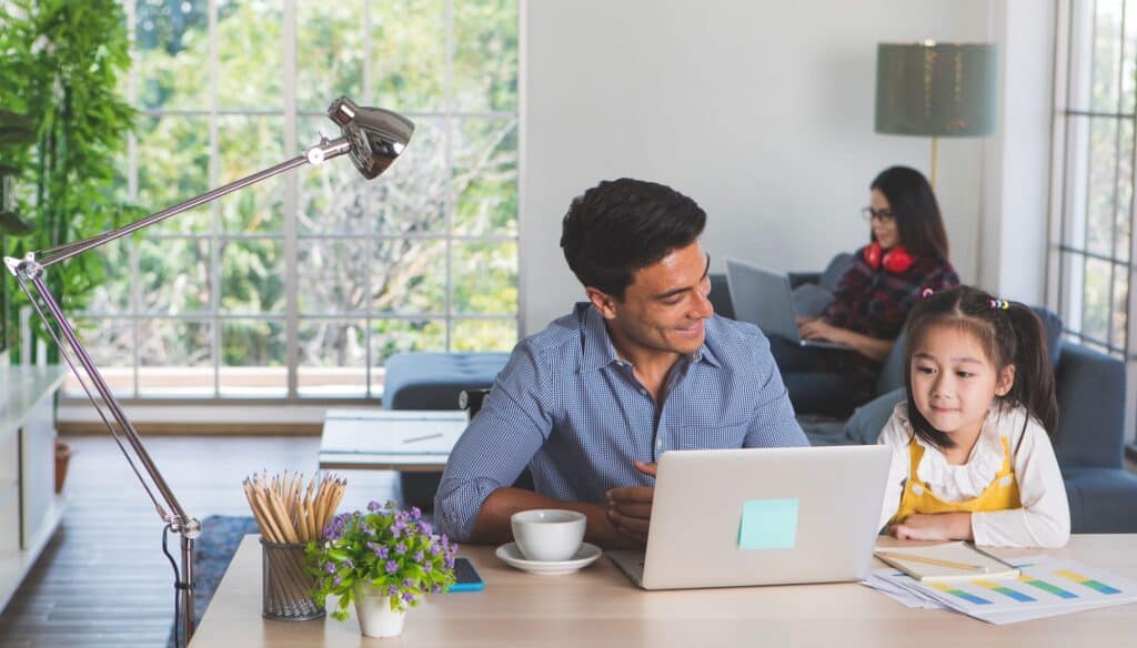A man works on a laptop at a desk with a young girl beside him, while a woman uses her laptop on the couch in the background, showcasing home efficiency and comfort while working at home.