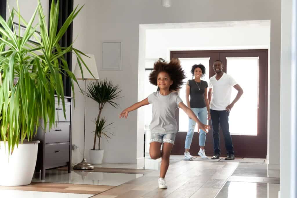 A young girl runs indoors with arms outstretched while two adults stand smiling near the doorway of a bright, modern home featuring potted plants and the best air conditioning for year-round comfort.