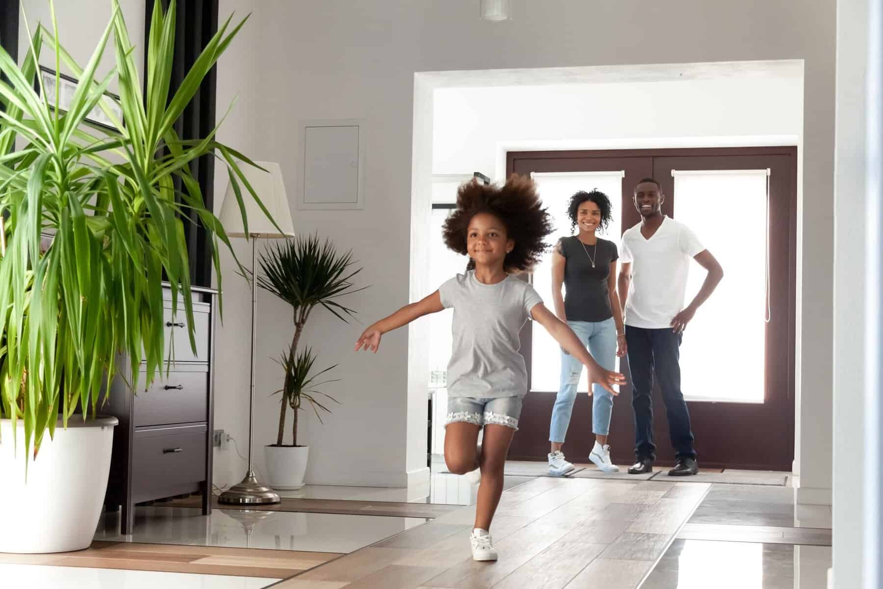 A young girl runs indoors with arms outstretched while two adults stand smiling near the doorway of a bright, modern home featuring potted plants and the best air conditioning for year-round comfort.