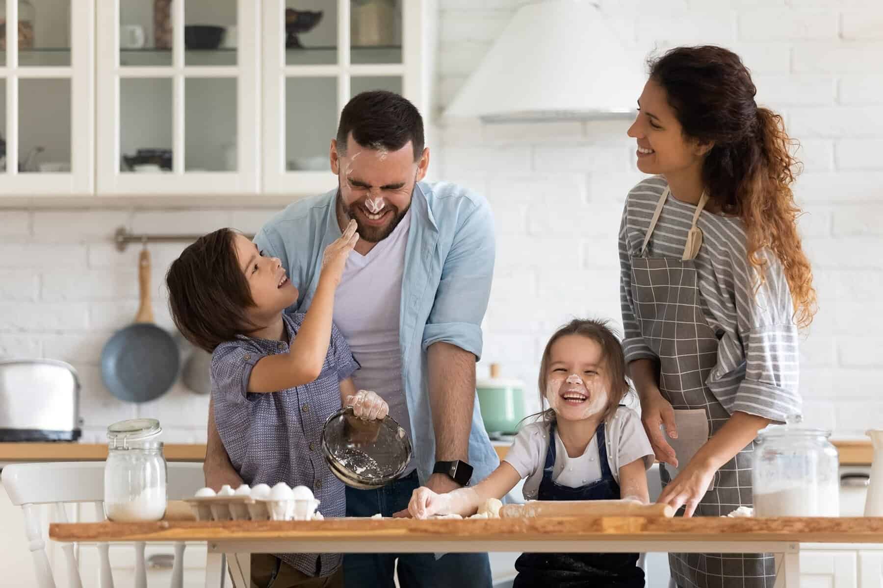 A family of four smiles and bakes together in the kitchen, flour everywhere. While a child playfully touches an adult's nose with flour, copper pipes gleam under the sink—no pinhole leak worries after their recent repair or replace decision.