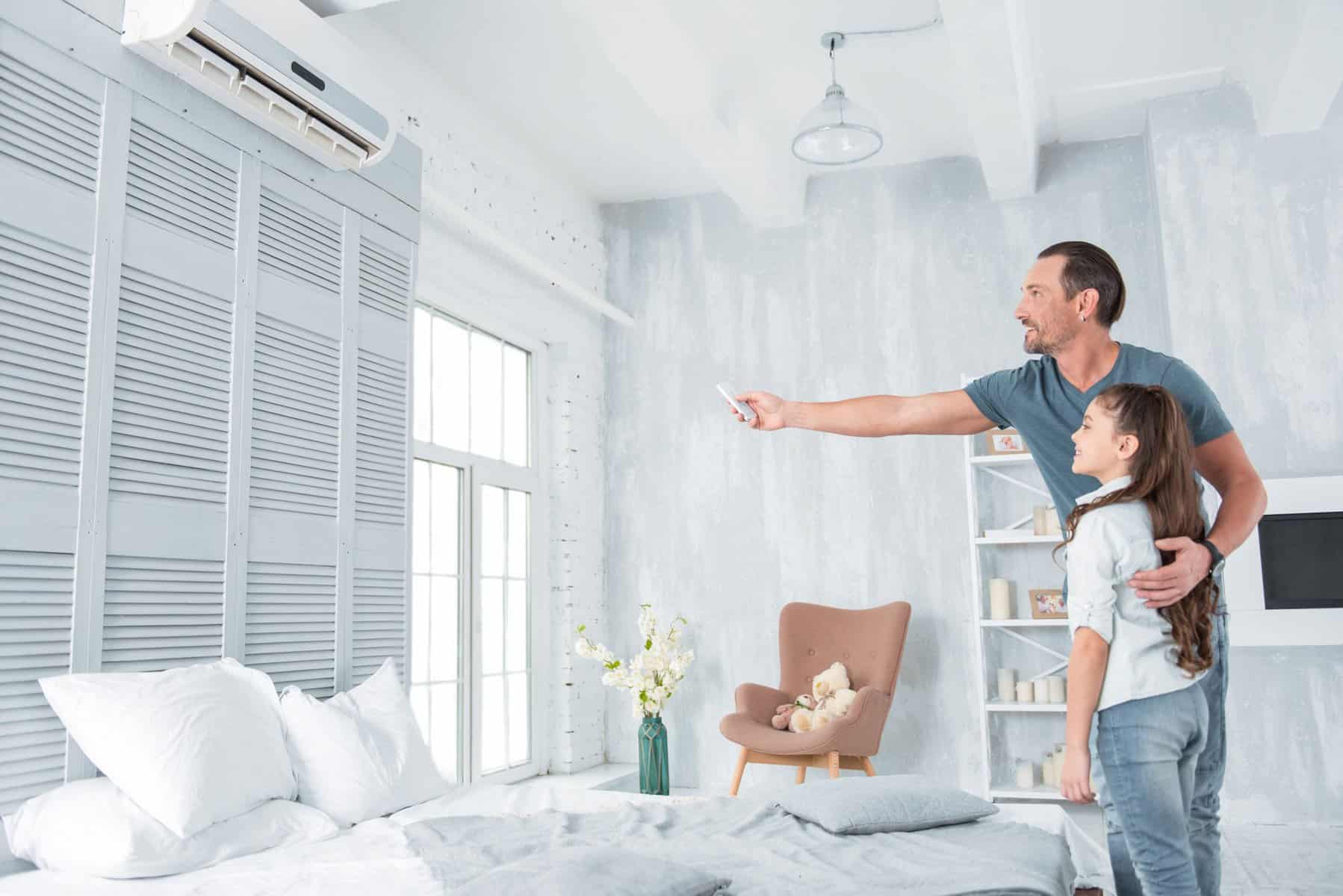 A man and a young girl stand in a bedroom, looking up as the man uses a remote to operate a mini split cooling system mounted on the wall.