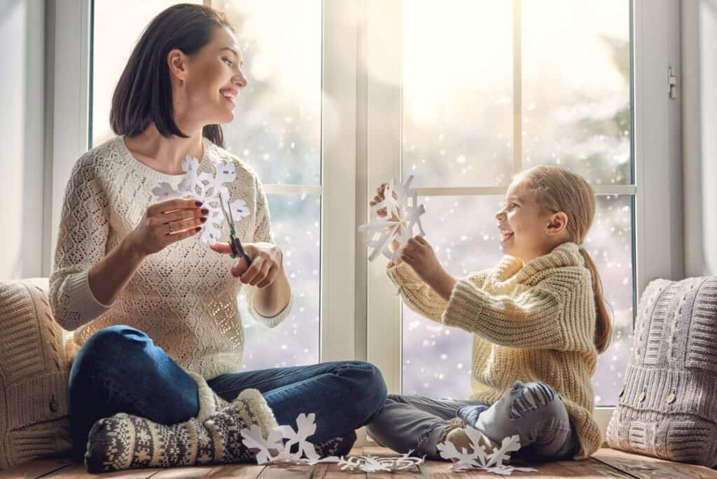 A woman and a young girl sit by a window, cutting and holding paper snowflakes, while their cozy home stays warm thanks to the heat pump system, with snow visible outside.