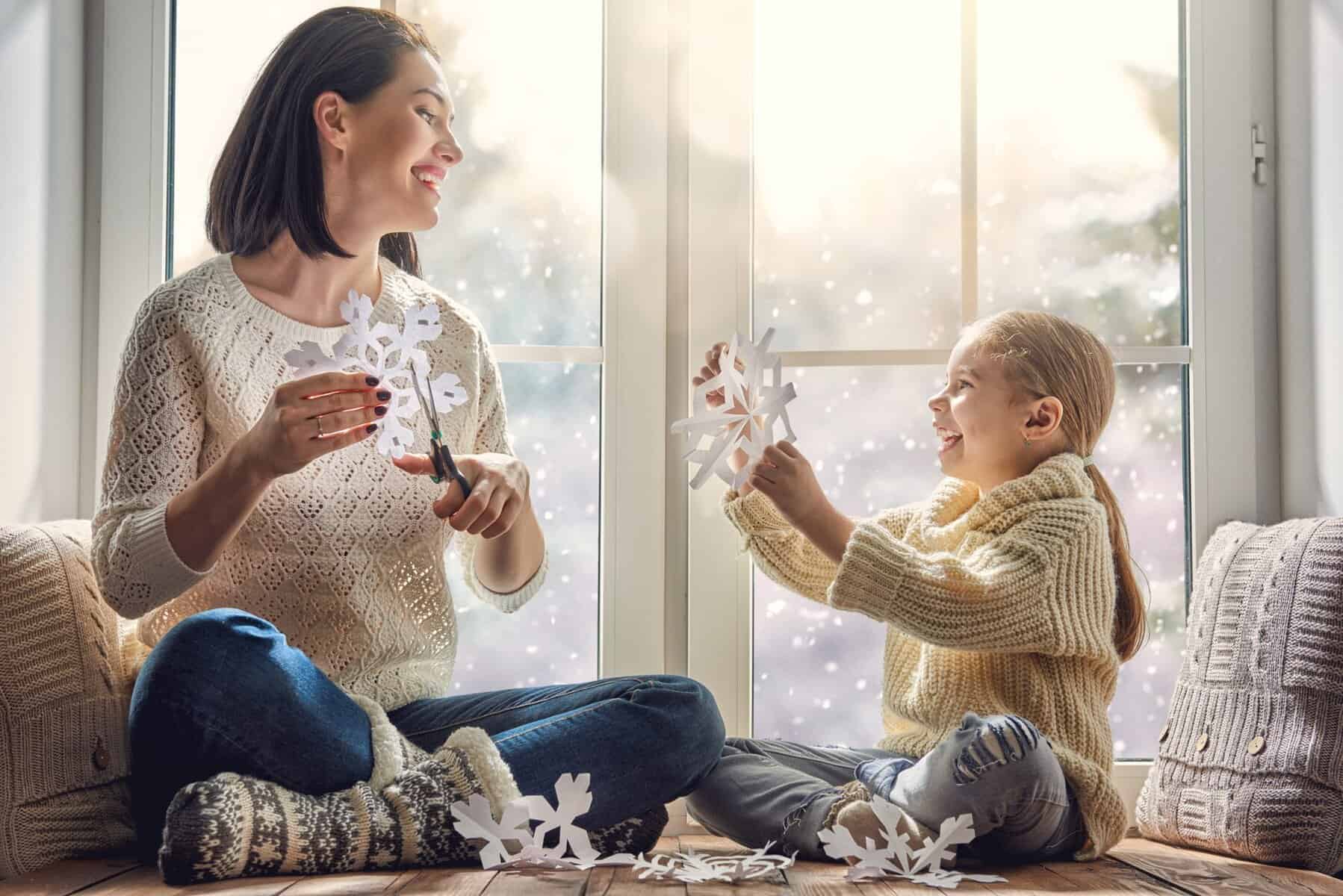 A woman and a young girl sit by a window, cutting and holding paper snowflakes, while their cozy home stays warm thanks to the heat pump system, with snow visible outside.