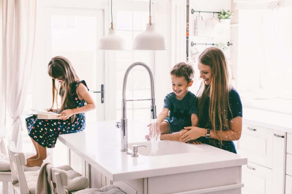 A woman and a young boy play with water at a kitchen island sink—a perfect time to remember handy Plumbing Maintenance Tips—while a girl sits nearby on a stool reading a book.