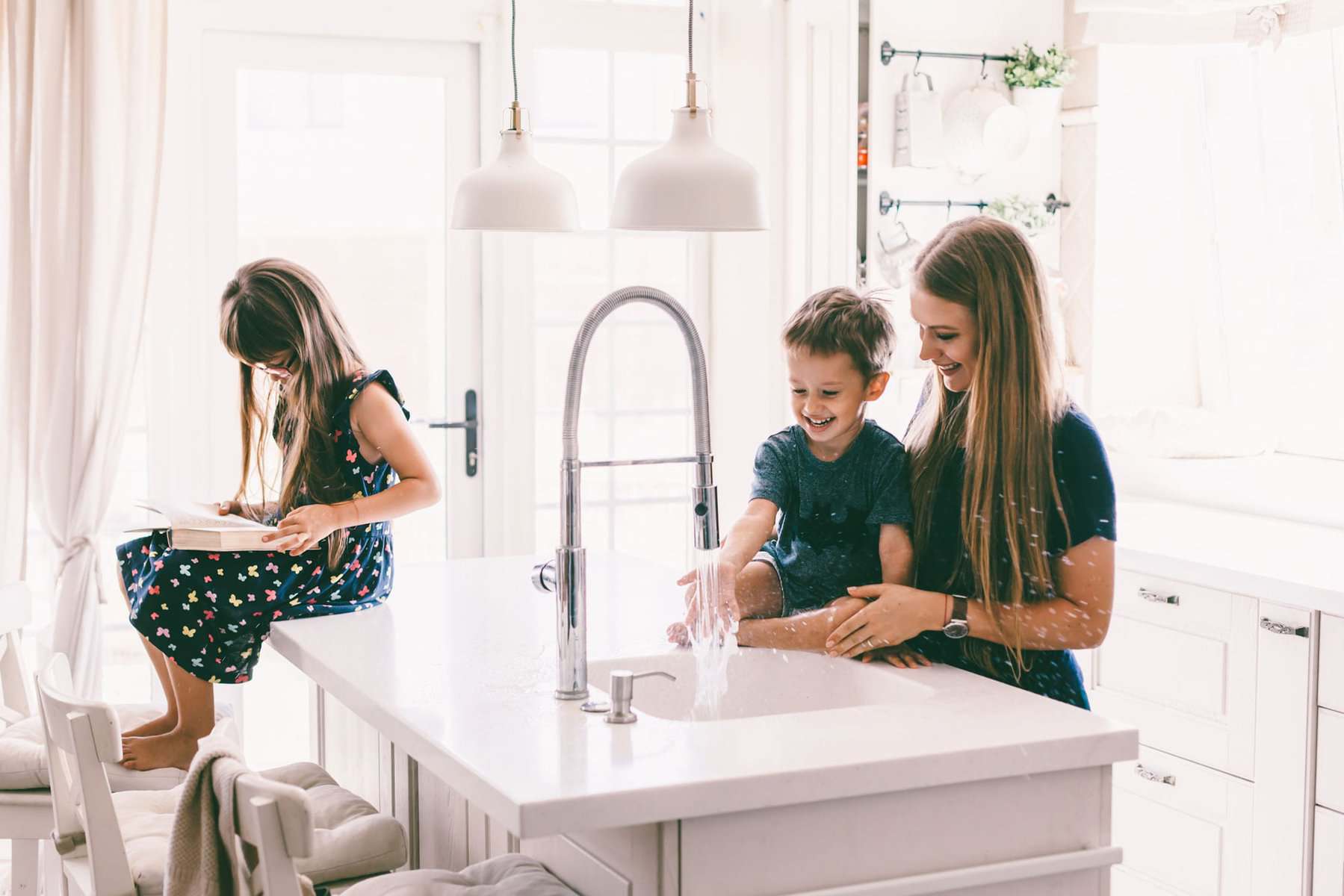 A woman and a young boy play with water at a kitchen island sink—a perfect time to remember handy Plumbing Maintenance Tips—while a girl sits nearby on a stool reading a book.