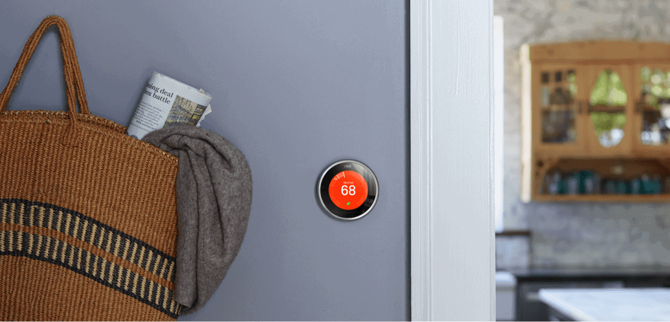 A Nest smart thermostat displaying 68 degrees is mounted on a gray wall next to a woven bag holding a rolled-up newspaper and scarf, showcasing the benefits of smart technology. A kitchen is visible in the background.