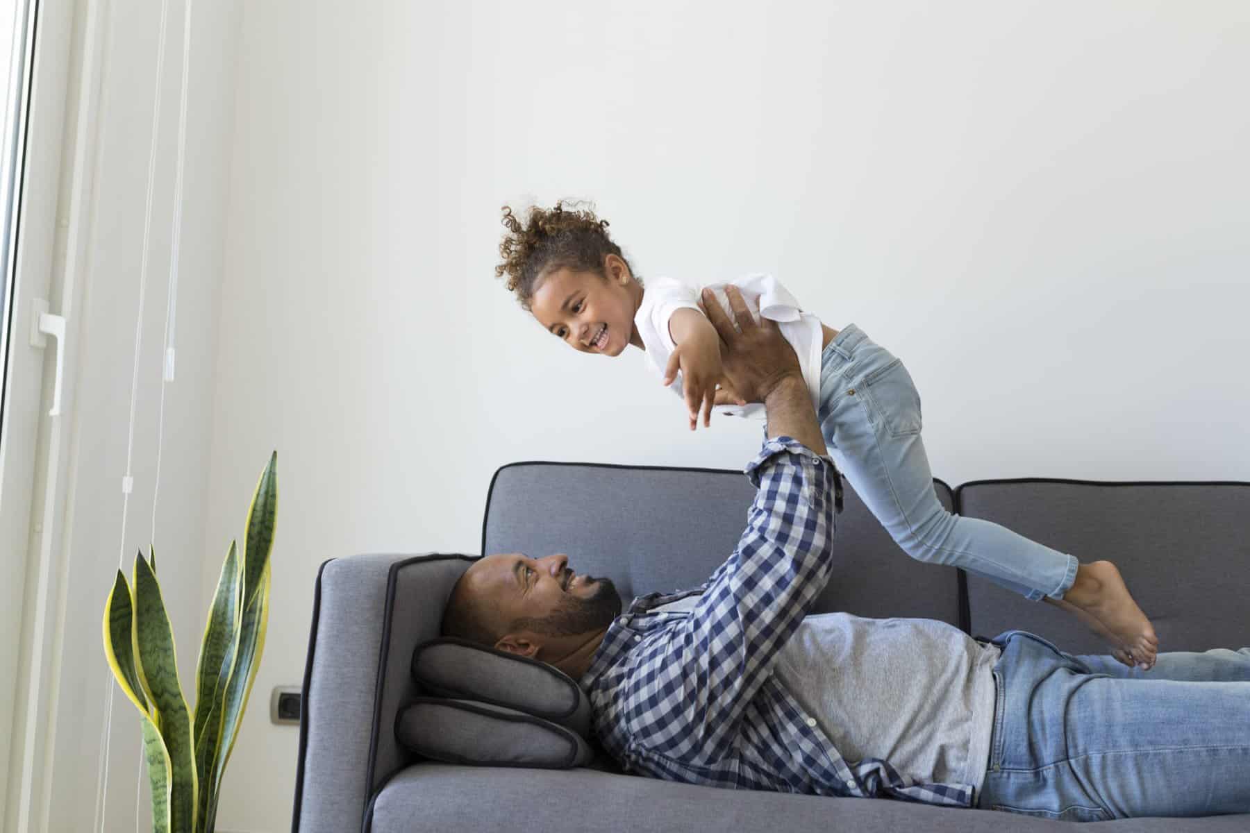 An adult lies on a sofa, lifting a smiling child above their chest near a potted plant, enjoying family time in the comfort of an efficient home heating and cooling system.