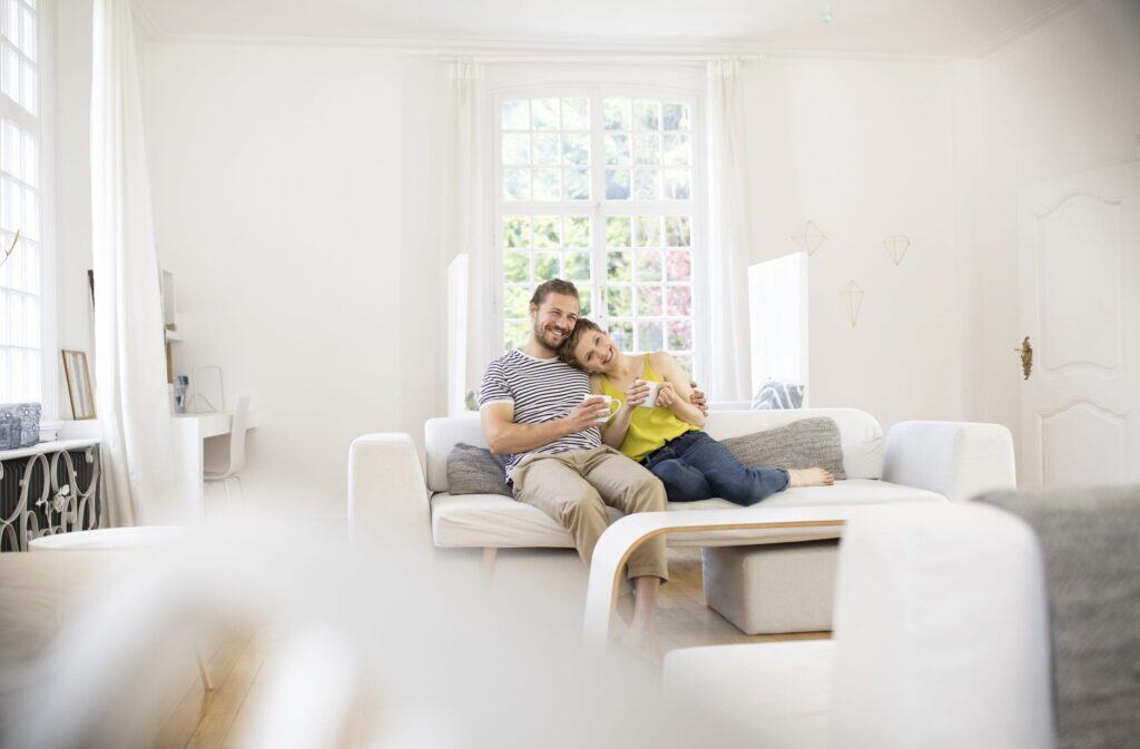 A man and a woman sit closely together on a white couch in a bright living room, smiling and holding mugs, discussing DIY heating maintenance to keep their home cozy.
