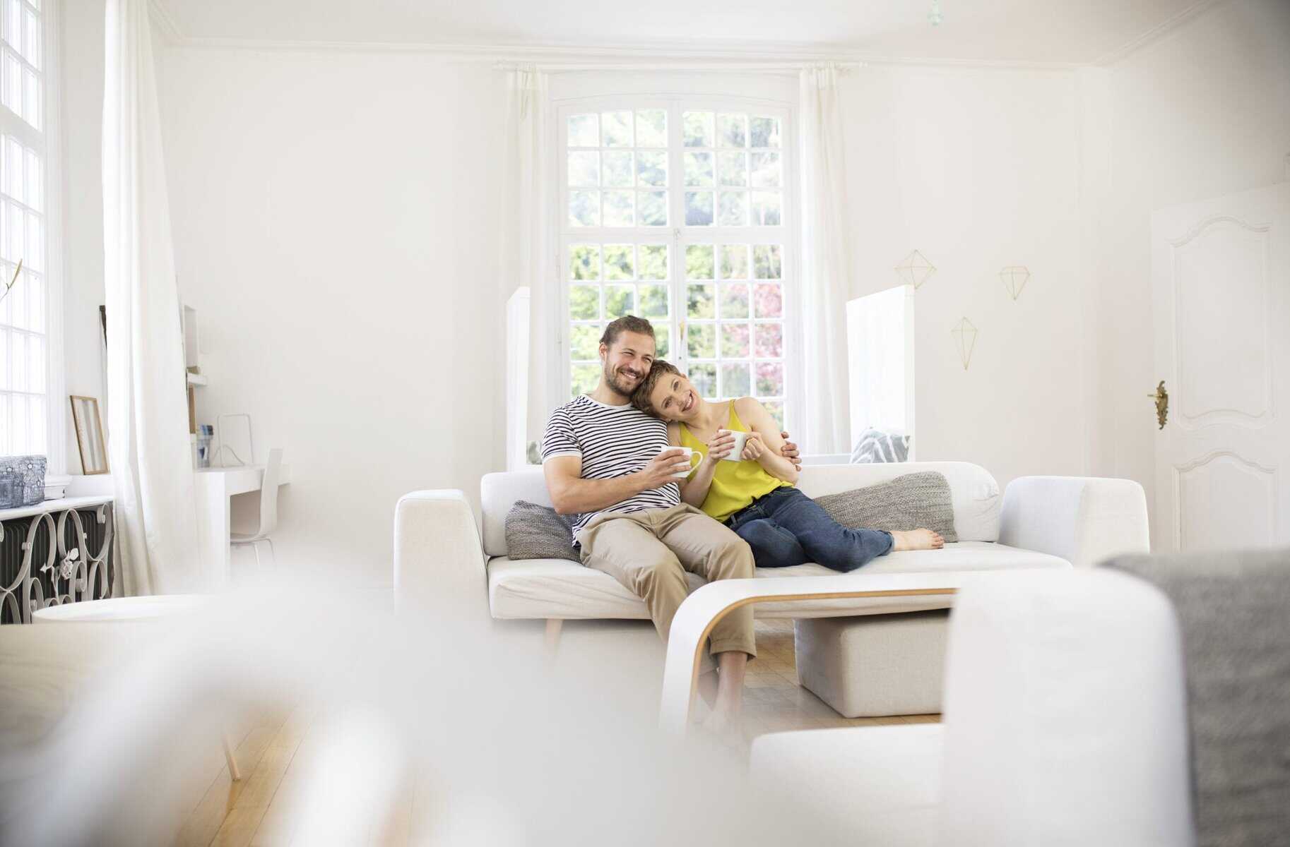 A man and a woman sit closely together on a white couch in a bright living room, smiling and holding mugs, discussing DIY heating maintenance to keep their home cozy.