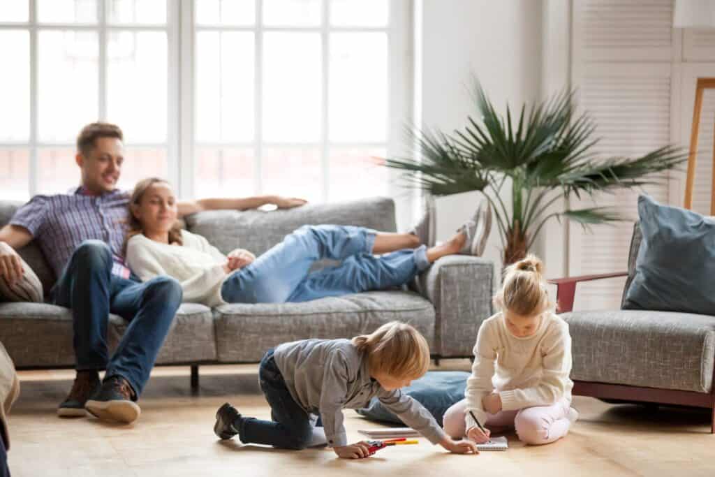 Two children draw with colored pencils on the floor while two adults, enjoying their relationship, sit on a sofa in the background of a bright living room with home humidifiers enhancing comfort.