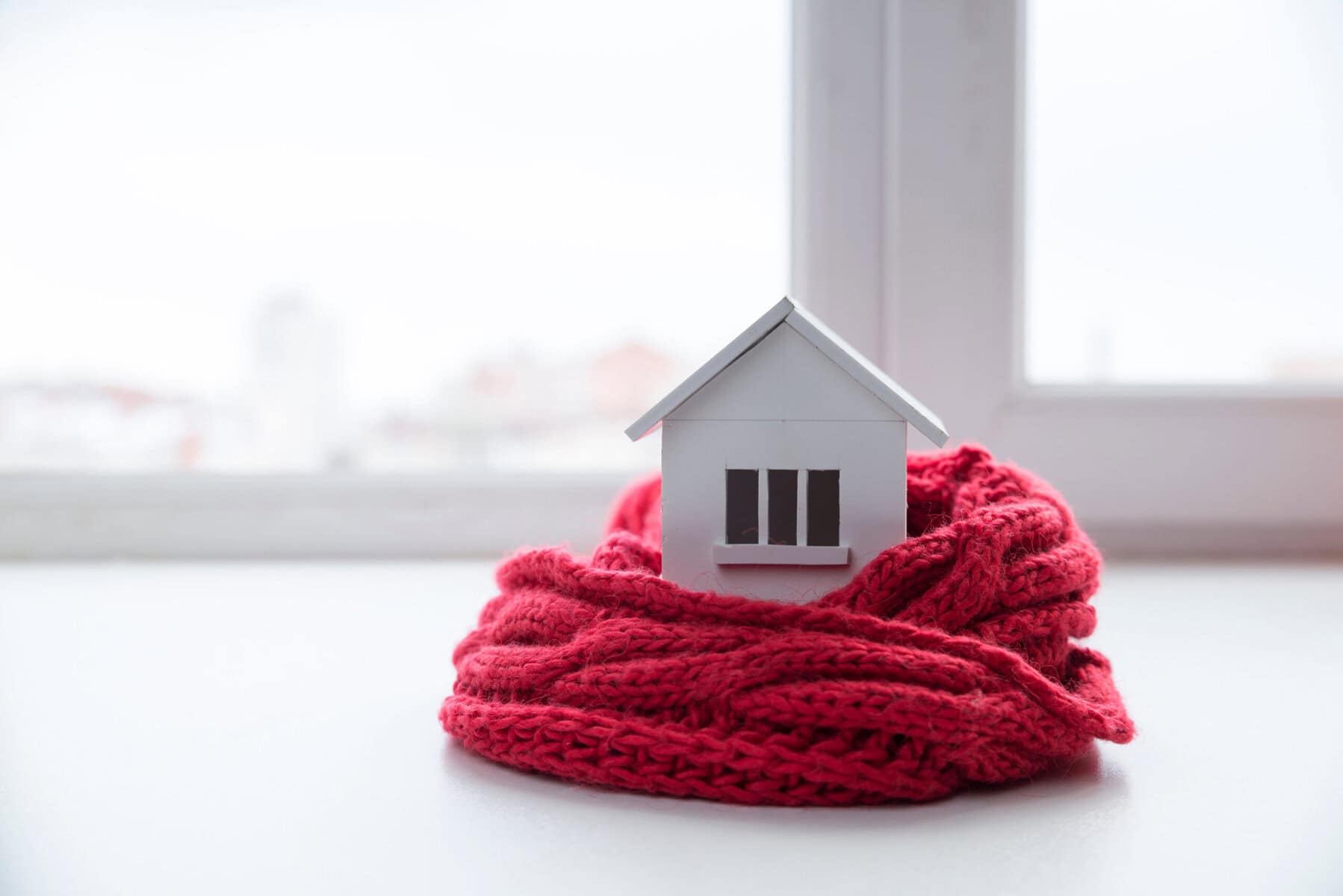 A small model house wrapped in a red knitted scarf sits on a white surface in front of a window, symbolizing an energy efficient home and offering inspiration for winter energy saving tips.