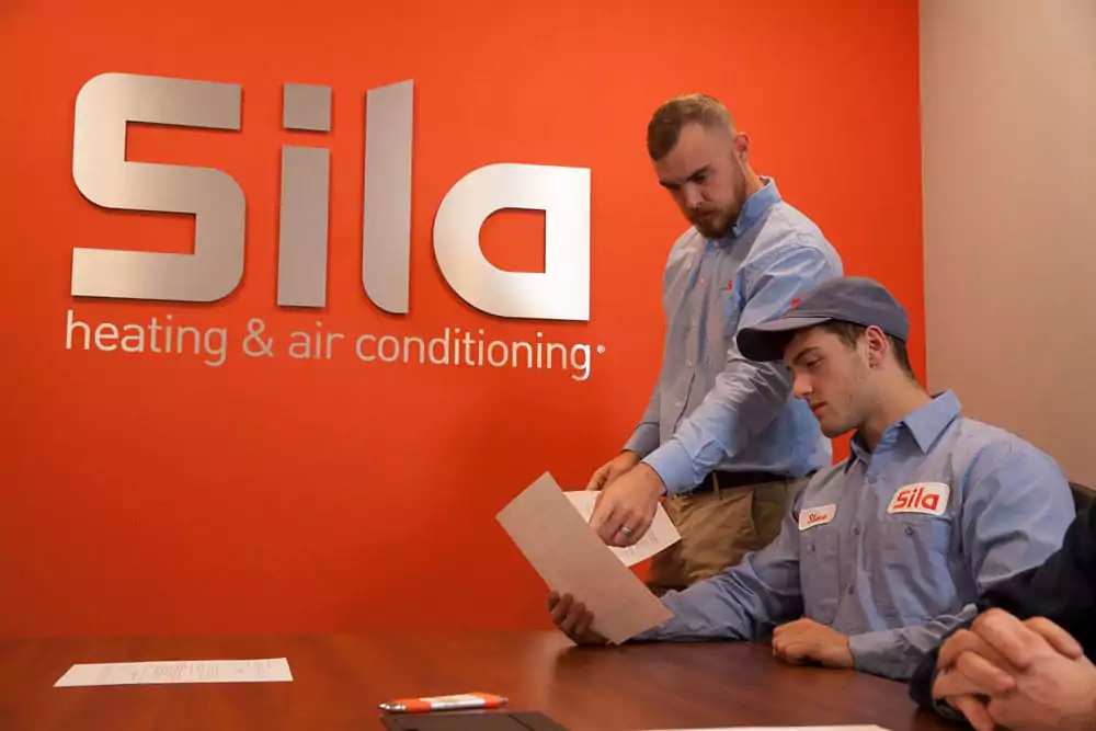Two employees in Sila uniforms review documents at a table in front of an orange wall with the Sila heating & air conditioning logo, offering a behind-the-scenes look at our team and what we're about.
