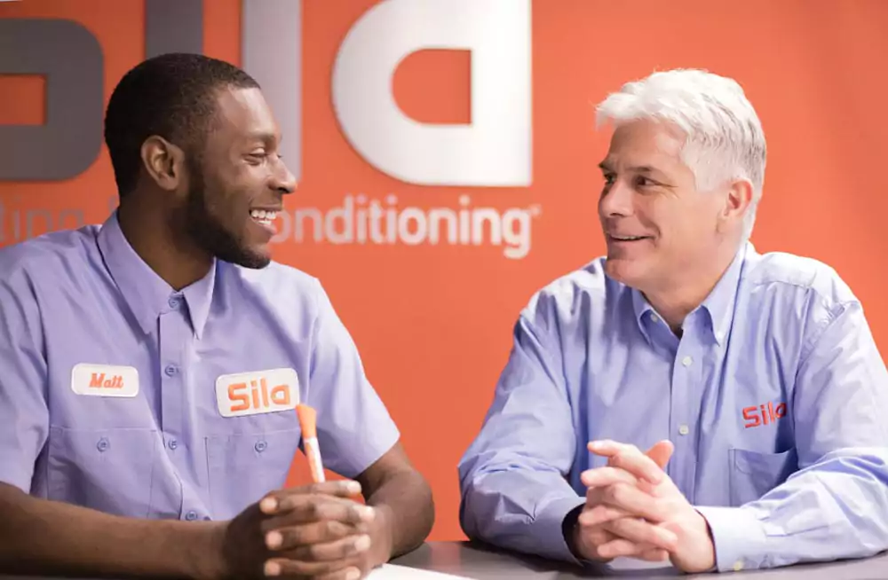 Two men in "Sila" uniforms sit at a table and smile at each other, discussing finance options, with an orange wall and company logo in the background.