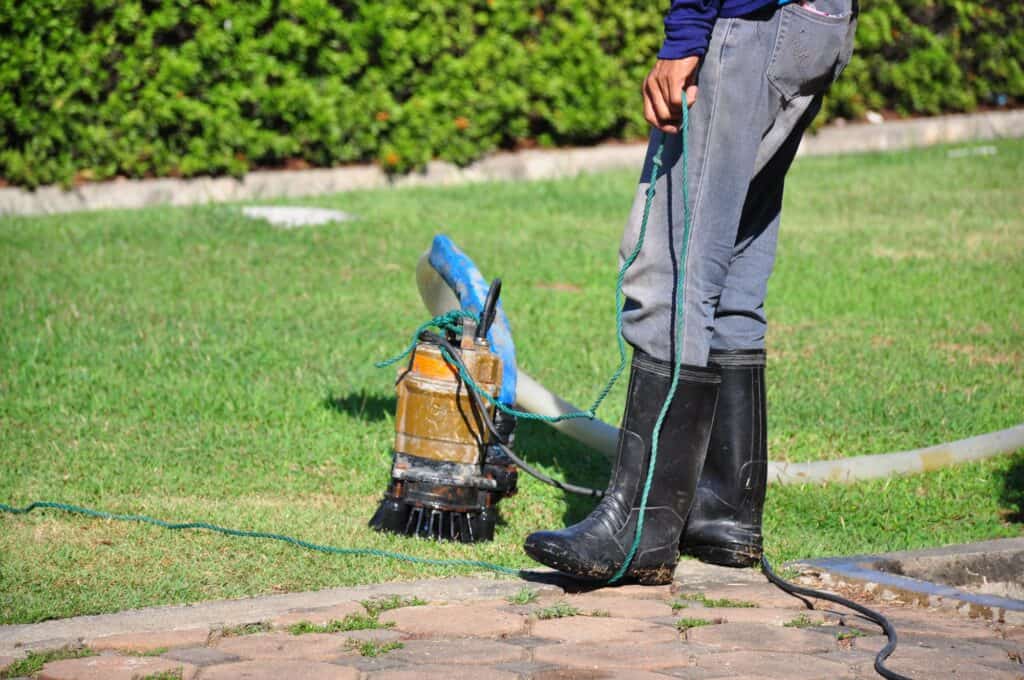 A homeowner practices sump pump maintenance, standing in rubber boots on grass while holding a rope. A submersible water pump and hose lie nearby, with shrubs in the background.