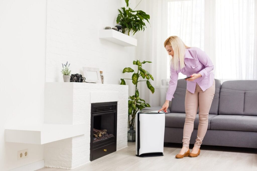 A woman in a light purple blouse presses a button on an air purifier with humidity control in a modern living room featuring plants, a fireplace, and a gray sofa.