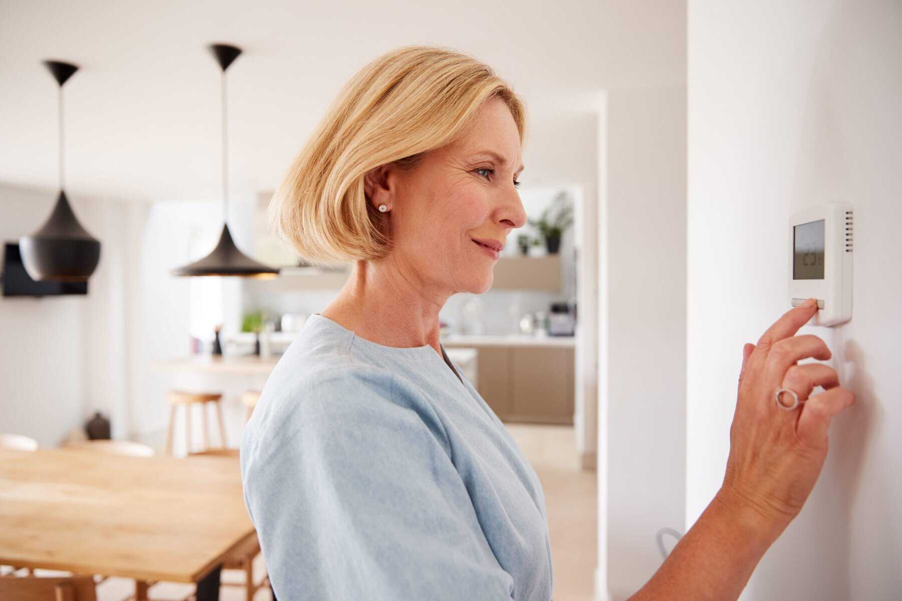 A woman stands in a modern kitchen adjusting a digital thermostat, staying alert to heating system signs that may indicate the need for heating repair.