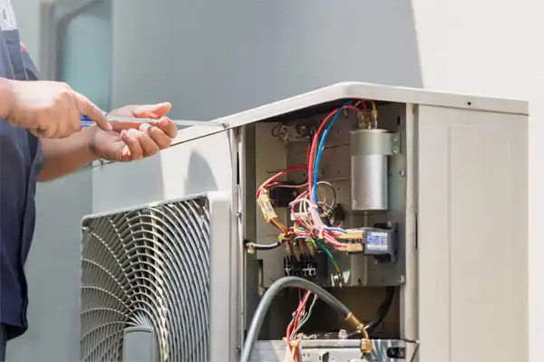 A person in Boston repairs or maintains an outdoor air conditioning unit with exposed wiring and parts using a screwdriver, showcasing essential skills for MA Furnace Repair.