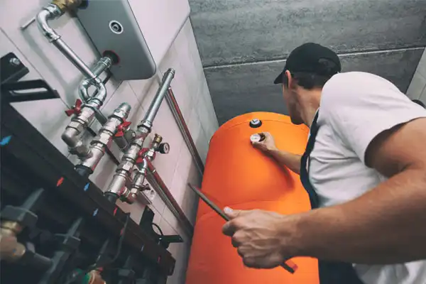 A worker inspects and adjusts a valve on a large orange tank in a utility room, performing general heating repair with visible pipes and equipment.