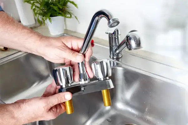 A person in Boston, MA holds a detached kitchen faucet with dual handles above a stainless steel sink, preparing for kitchen plumbing installation or replacement.
