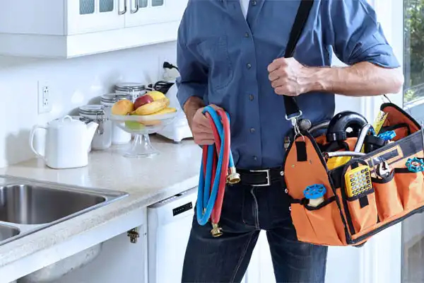 A person holding a tool bag and flexible hoses stands in a kitchen near a sink, suggesting Kitchen Plumbing Repair in Boston, MA.