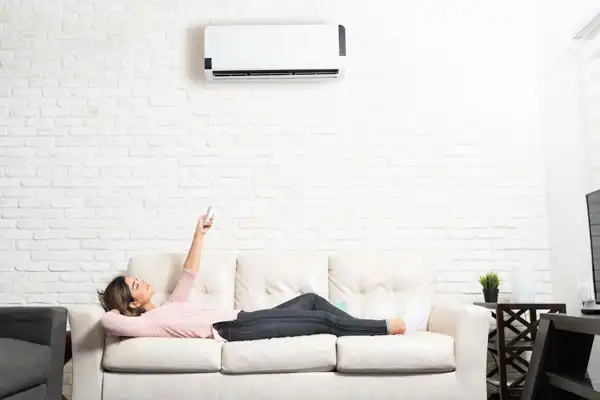 A woman lies on a beige sofa, pointing a remote control at a wall-mounted air conditioner after a recent mini-split installation in her bright Boston, MA living room.
