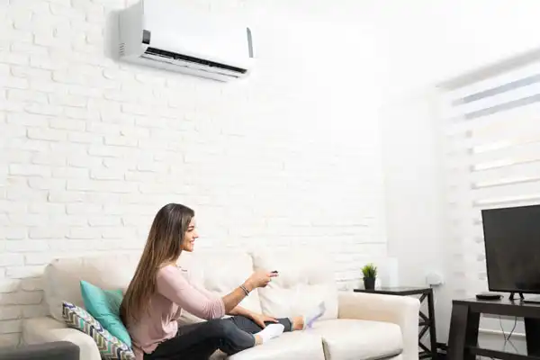 A woman sits on a couch in her Boston home, holding a remote control and watching TV, with an air conditioner above her—reminding homeowners of the importance of regular Mini-Split Maintenance in MA.