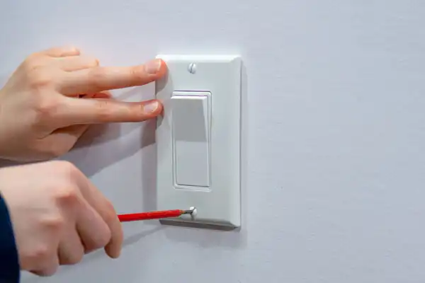 A person uses a screwdriver to remove or tighten a screw on a white electrical switch plate mounted on a wall, part of routine Electrical Switches maintenance in Boston MA.
