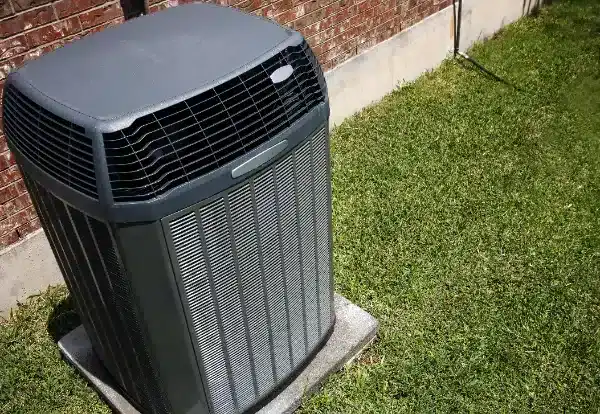A central air conditioning unit, expertly installed on a concrete pad next to a brick wall, sits amid lush green grass—showcasing professional air conditioning installation in Boston, MA.
