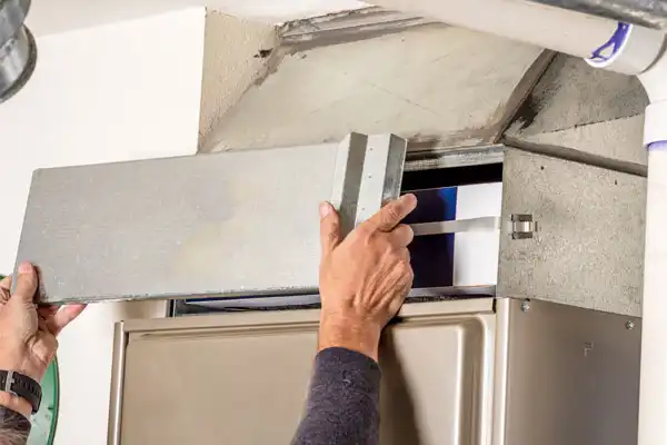 A person performs furnace maintenance in Boston, MA, installing or removing a filter from a residential HVAC unit while holding the metal cover panel.