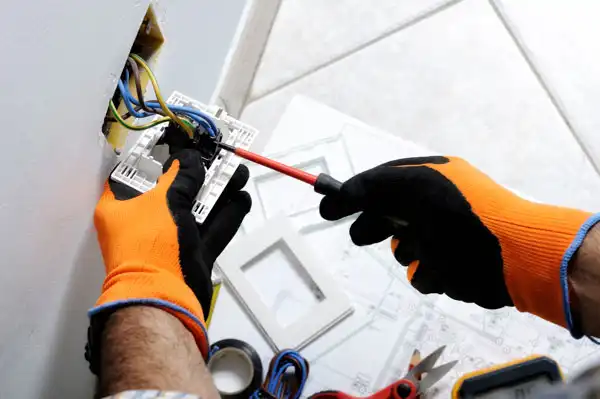A person in Boston, MA wearing orange and black gloves uses a screwdriver to perform general electrical repair on wiring in a wall outlet. Tools and electrical tape are visible nearby.