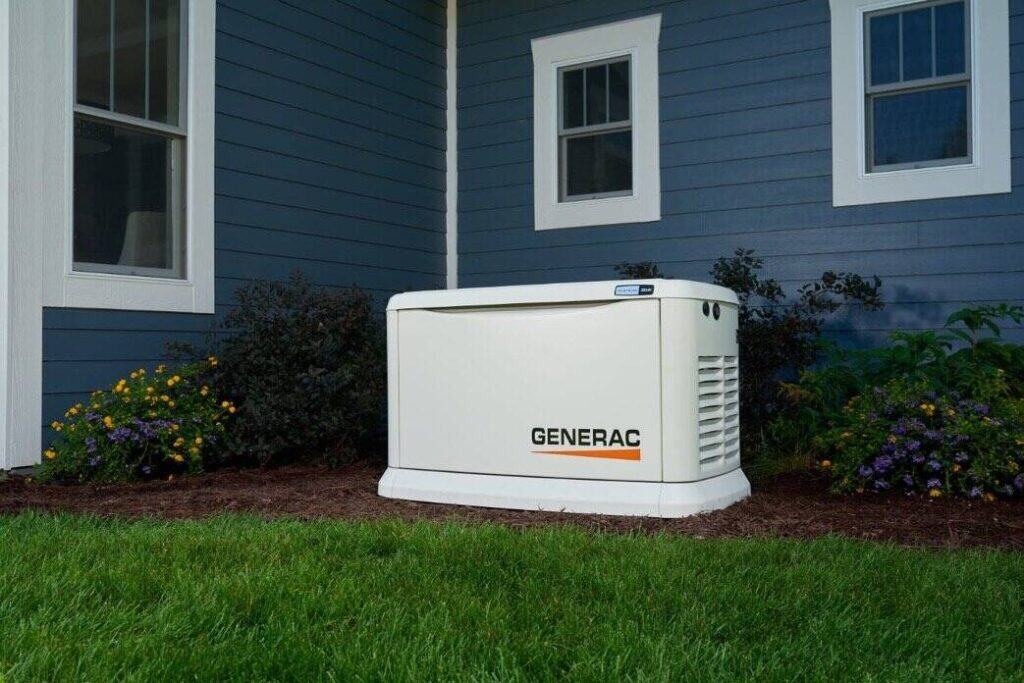 A Generac standby generator is installed outside a house with blue siding and white trim, surrounded by mulch and plants.