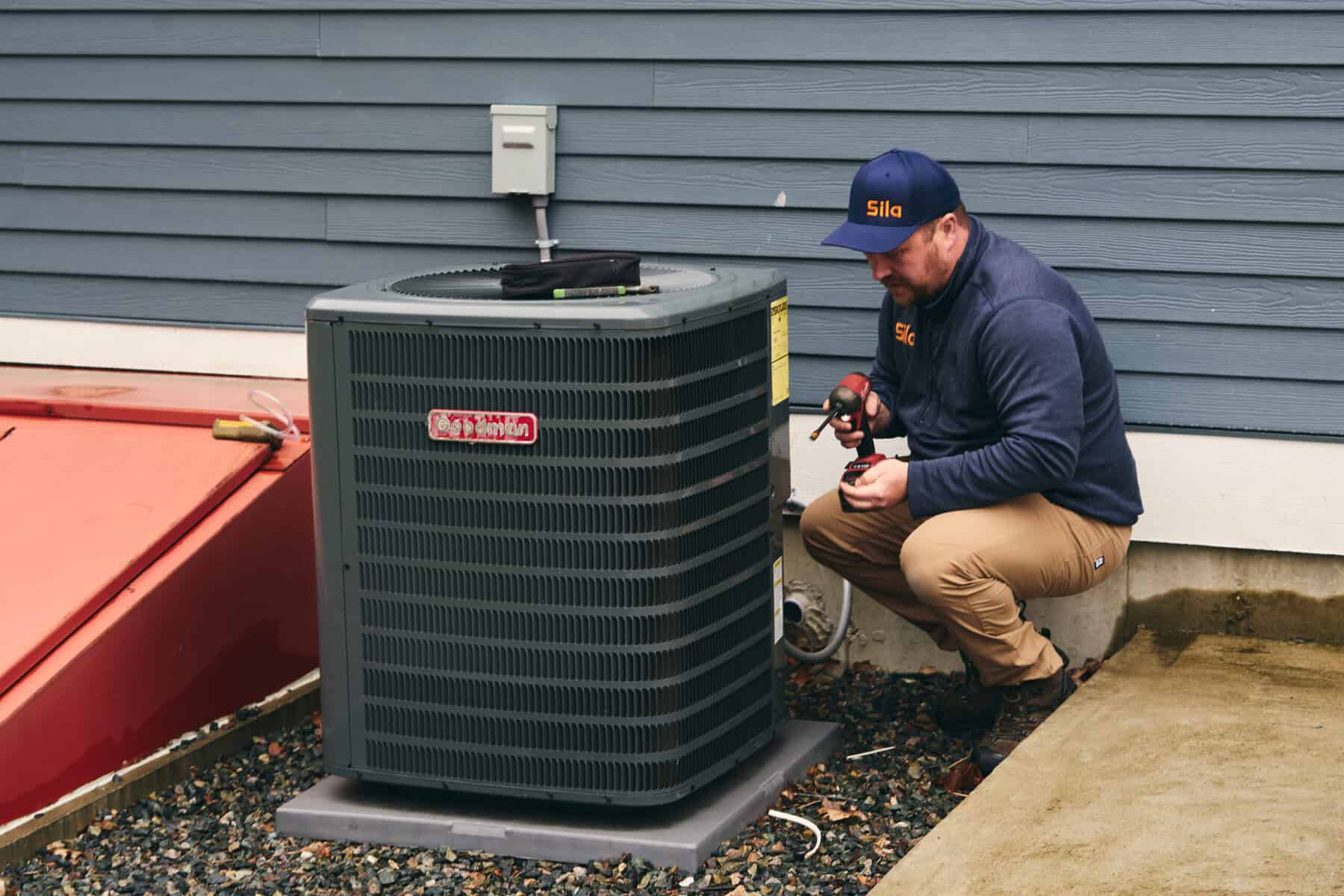 A technician in a navy jacket and cap is servicing an outdoor air conditioning unit next to a house with blue siding, showcasing the expertise found in Plumber Repair Boston MA professionals.