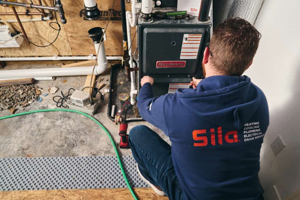 A technician in a blue hoodie kneels while servicing a Goodman furnace in a utility room, with various pipes and tools visible—your trusted choice for plumber repair in Boston, MA.