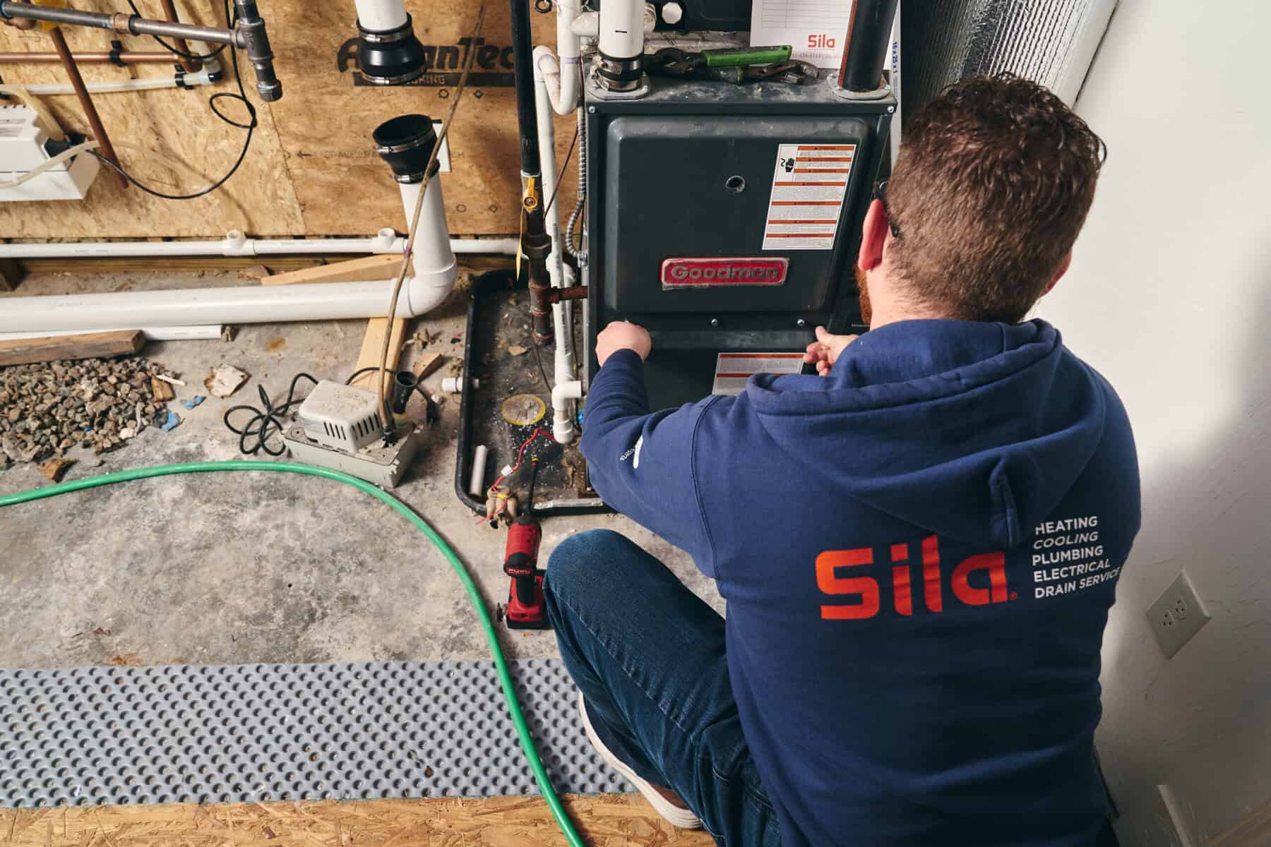 A technician in a blue hoodie kneels while servicing a Goodman furnace in a utility room, with various pipes and tools visible—your trusted choice for plumber repair in Boston, MA.