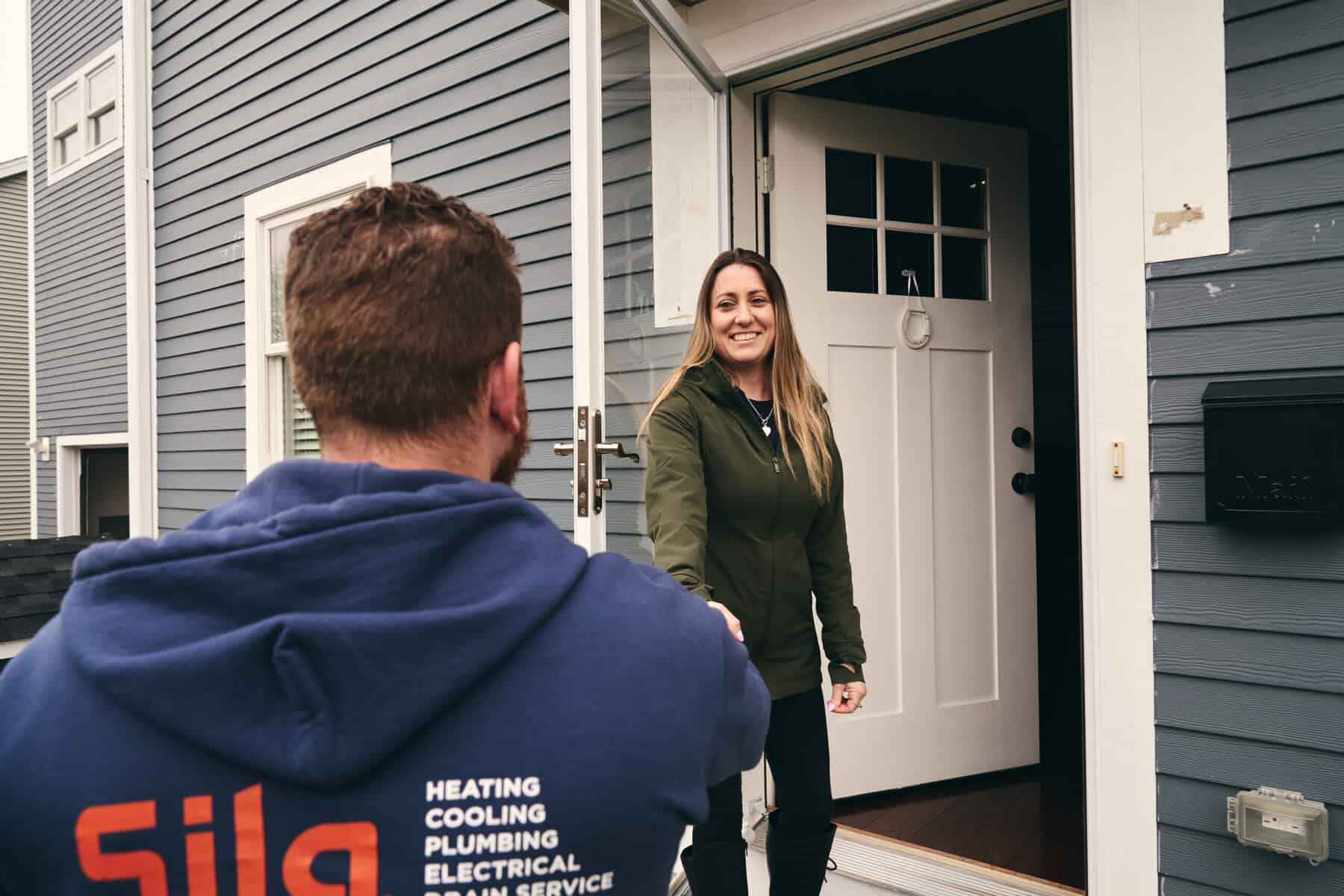 A woman stands at her front door smiling and shaking hands with a man in a blue hoodie featuring heating, cooling, plumbing, electrical services, and whole home generators Boston text on the back.