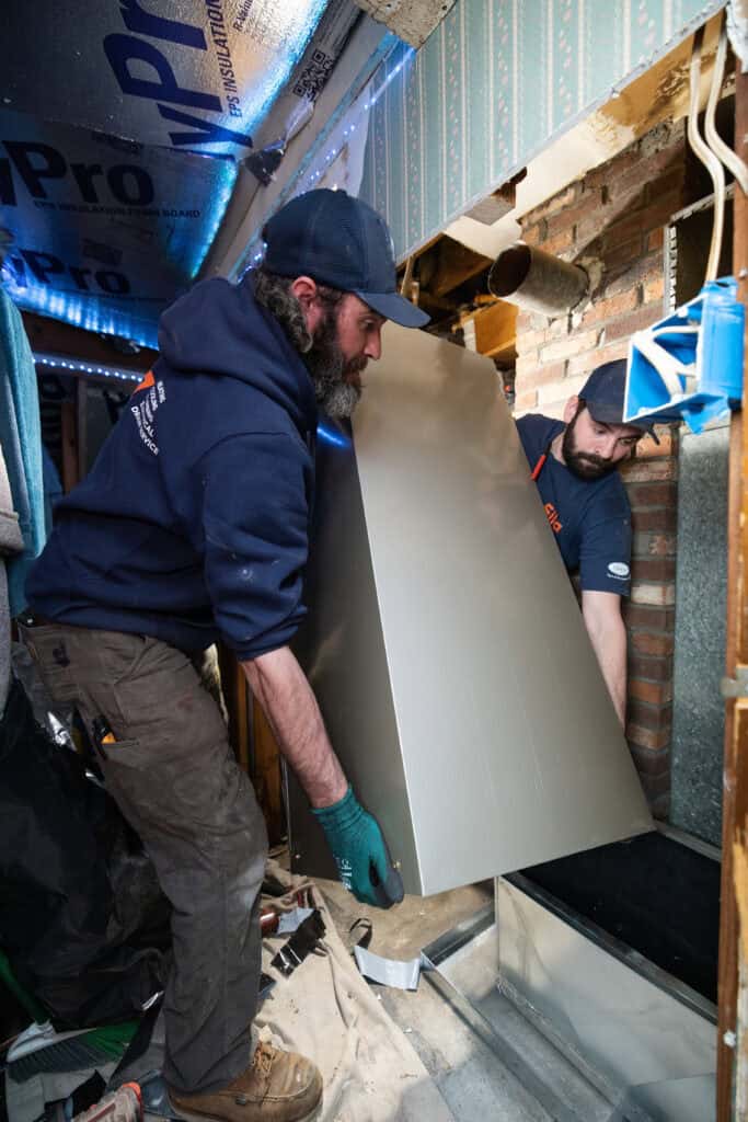 Two men install a large metal panel inside a partially constructed wall, surrounded by tools and construction materials.