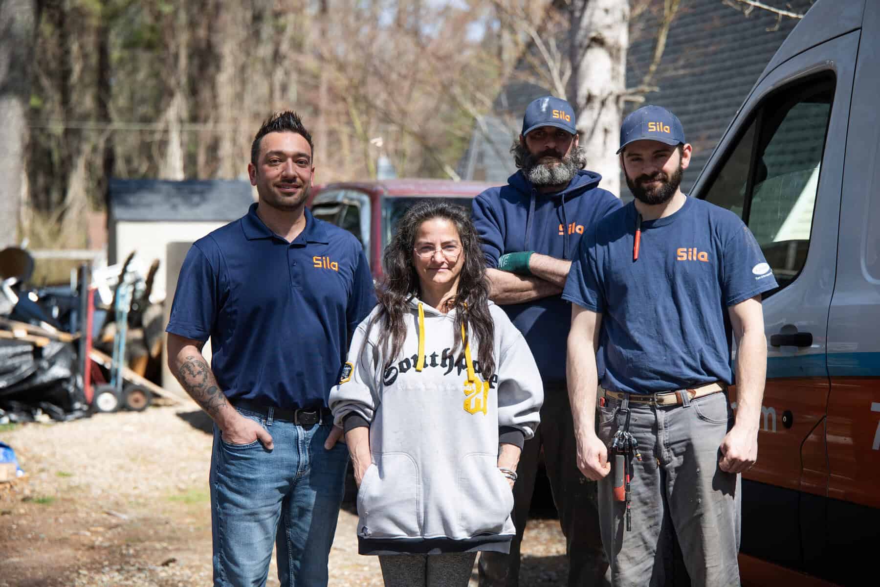 Four adults stand outdoors near a van and house, three wearing "Sila" uniforms and one in a gray sweatshirt, posing for a group photo on a sunny day during the Power the Nation Giveaway.