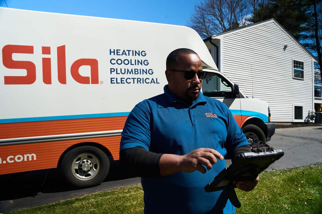 A worker in a blue Sila uniform uses a tablet in front of a Sila service van parked outside a house.