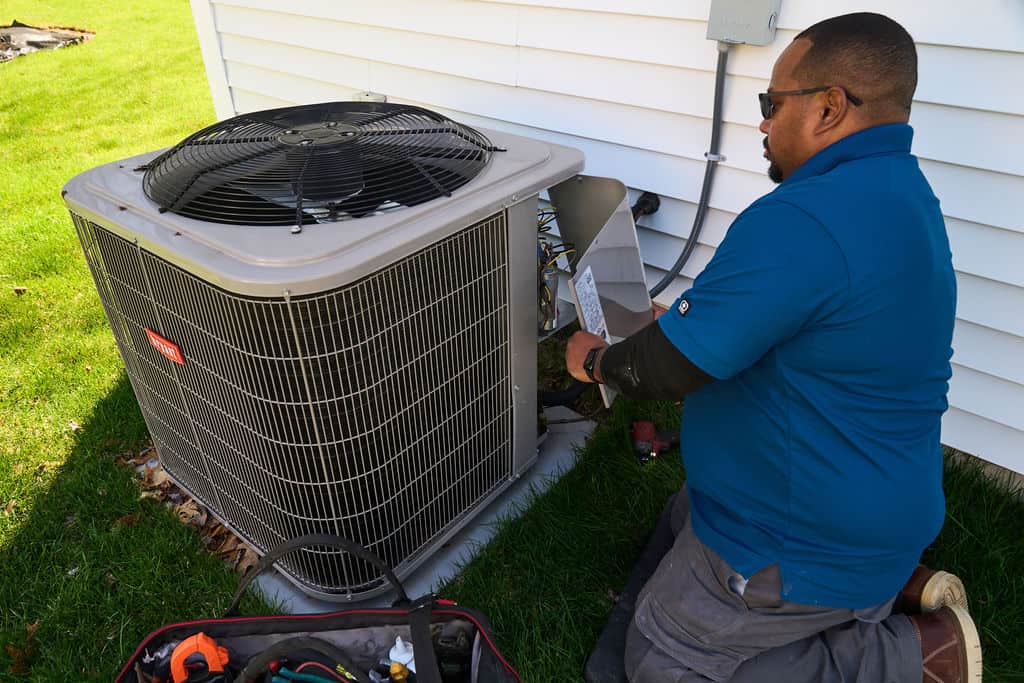 A technician in a blue shirt repairs or inspects an outdoor air conditioning unit next to a house in Winchester, MA, with tools visible on the ground nearby.