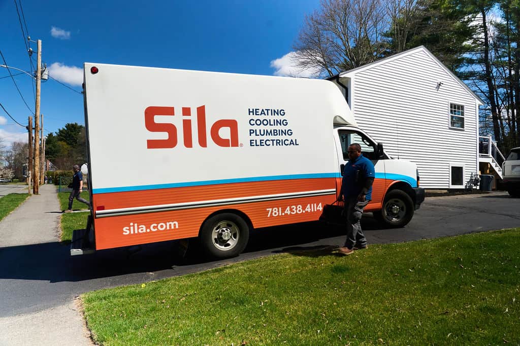 A Sila service van parked in a residential driveway, with a person standing beside it. The van advertises heating installation MA, as well as cooling, plumbing, and electrical services.