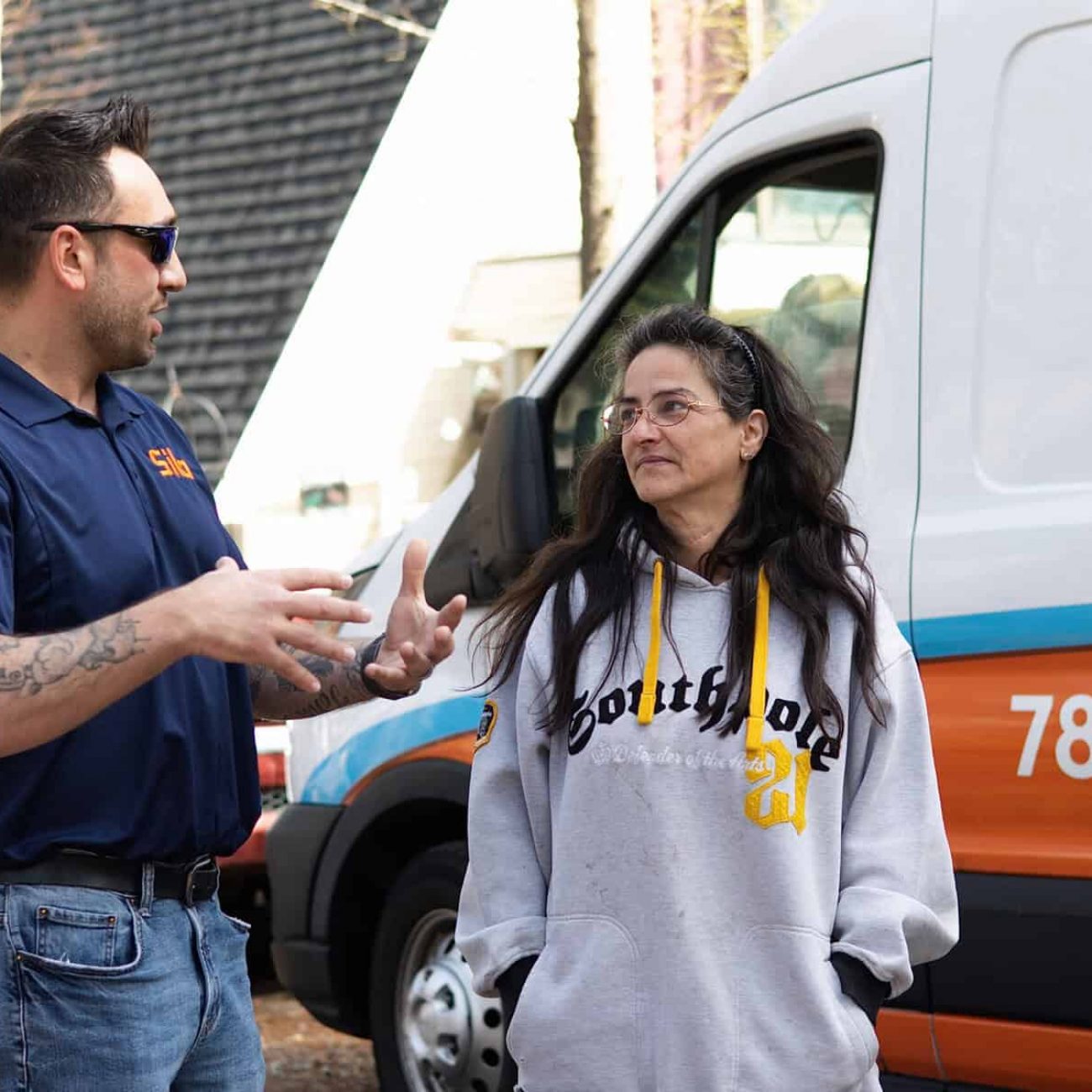 A man in a blue shirt talks to a woman in a gray hoodie near a van with orange and blue lettering and a phone number on the side.