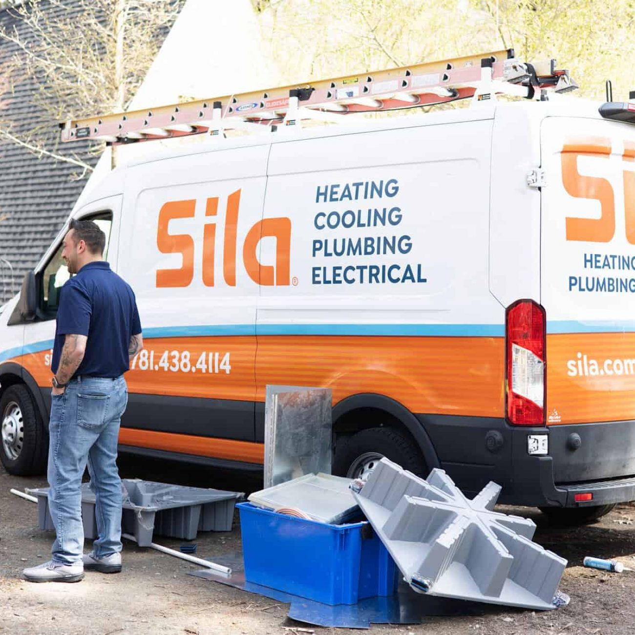 A plumber and a woman talk next to a Sila service van with heating, cooling, plumbing, and electrical services listed; tools and equipment are on the ground nearby.
