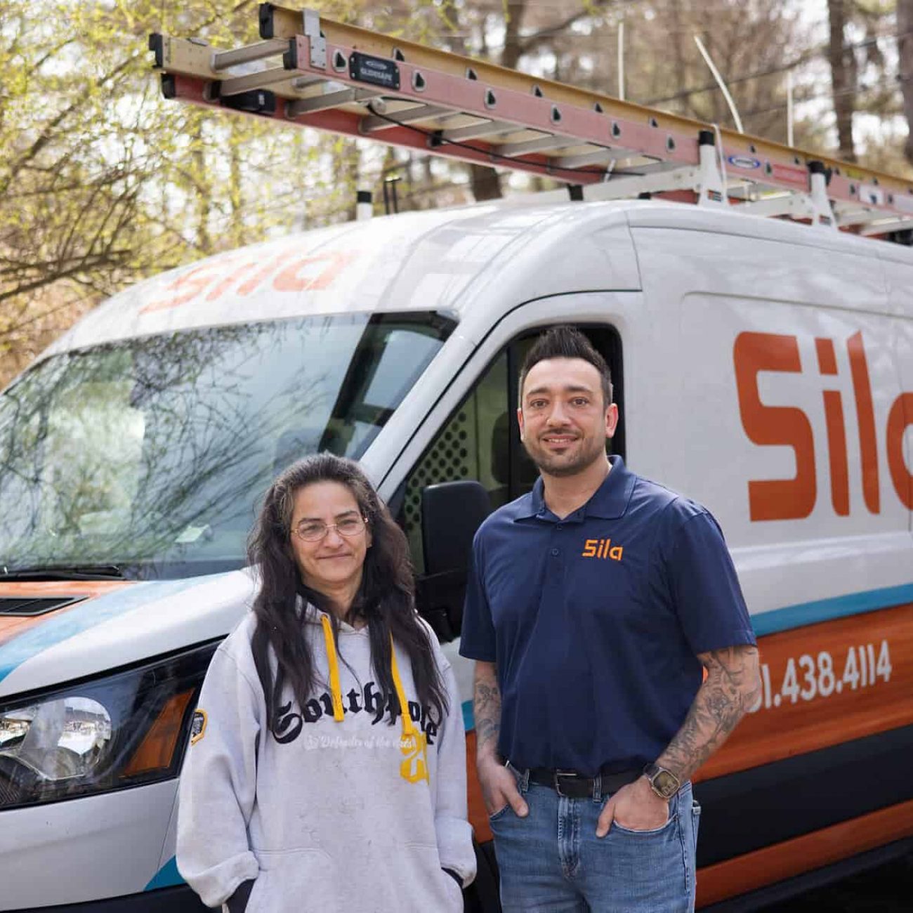 Two people stand in front of a Sila service van with a ladder on top, branded with "Heating, Cooling, Plumbing, Electrical," parked outdoors near trees in Cambridge.