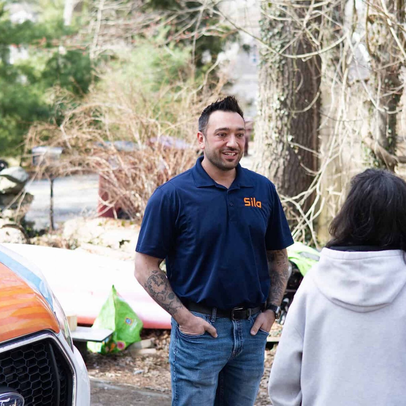 A man in a blue Sila polo shirt stands outdoors talking to a woman in a light hoodie near a white and orange vehicle, with trees and yard items in the background.